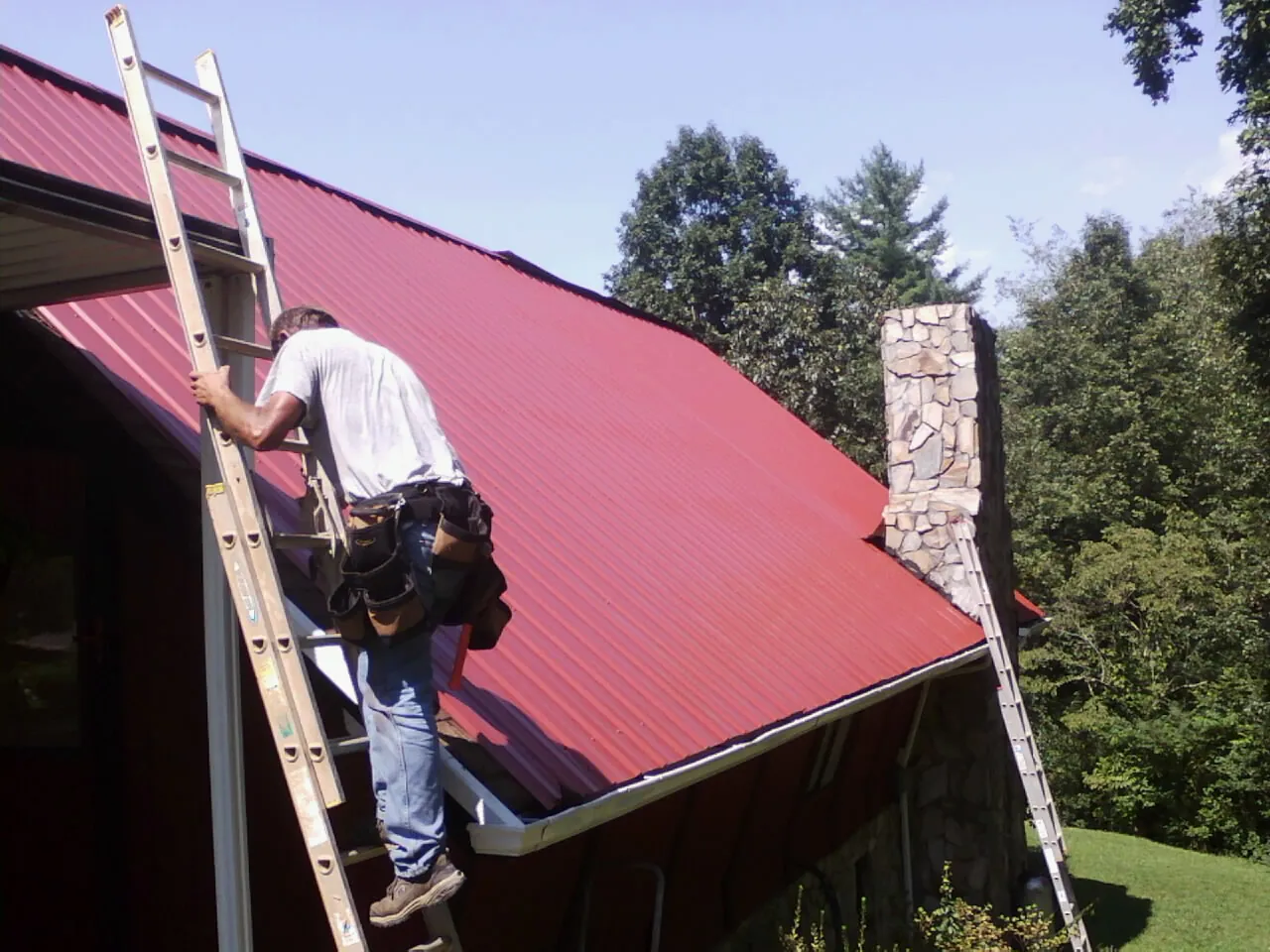 Roofer on a ladder beside a red metal roof and stone chimney — Soffit & Fascia Repair by Roofing Brothers in Ridgeville